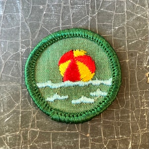 Puede incluir: Un parche de Girl Scout bordado en verde y blanco con una pelota de playa roja y amarilla flotando en agua azul.