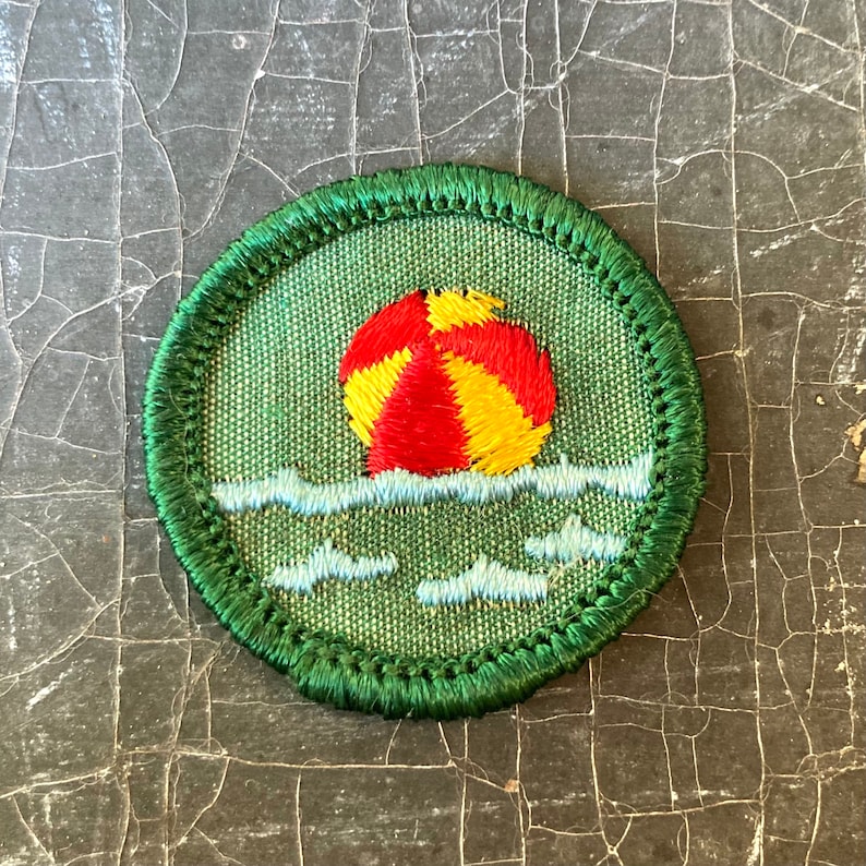 Puede incluir: Un parche de Girl Scout bordado en verde y blanco con una pelota de playa roja y amarilla flotando en agua azul.