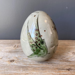 Puede incluir: Un jarrón de cerámica con forma de huevo con un pájaro verde pintado en él. El pájaro tiene alas marrones y verdes y un ojo marrón. El jarrón está decorado con motas marrones y un esmalte gris claro.