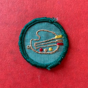 May include: A green and brown embroidered patch with a paint palette design. The palette has red, yellow, and brown paint on it.