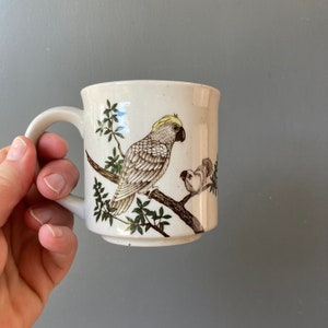 May include: A white ceramic mug with a brown and green design of two birds. The larger bird is a cockatoo with a yellow crest. The smaller bird is perched on a branch.