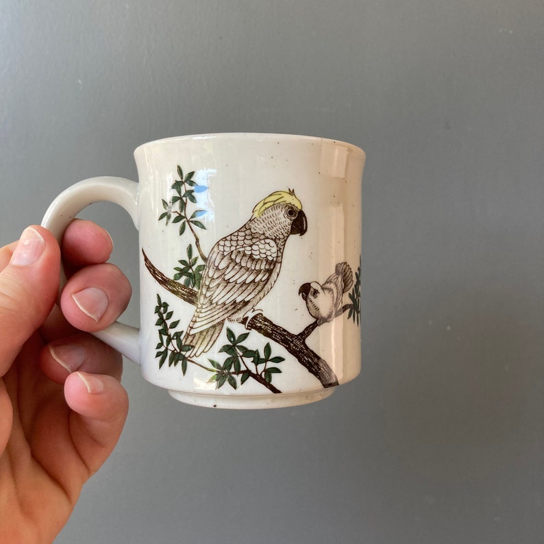 May include: A white ceramic mug with a brown and green design of two birds. The larger bird is a cockatoo with a yellow crest. The smaller bird is perched on a branch.