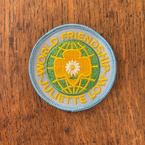 May include: A round patch with the words "World Friendship Juliette Low" in yellow text. The patch features a green and yellow design with a white flower in the center, all set against a light blue background.