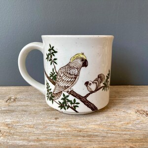 May include: White ceramic mug with a brown and green illustration of two parrots on a branch. The larger parrot has a yellow crest.