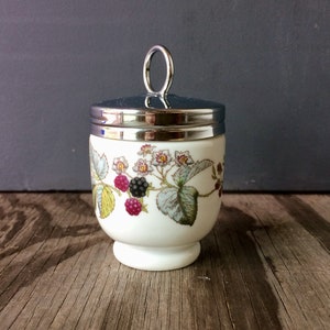 Op de afbeelding: Een wit keramiek eierdopje met een zilveren deksel en een bloemenontwerp met bramen en bloemen. Het eierdopje heeft een ronde basis en een zilveren handvat op het deksel.