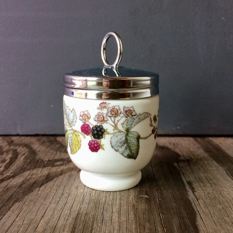 Op de afbeelding: Een wit keramiek eierdopje met een zilveren deksel en een bloemenontwerp met bramen en bloemen. Het eierdopje heeft een ronde basis en een zilveren handvat op het deksel.