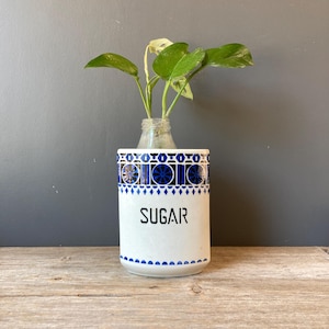 May include: White ceramic sugar container with blue geometric patterns and the word "SUGAR" in black. A small glass bottle holds a green plant, placed inside the container. The container sits on a wooden surface.