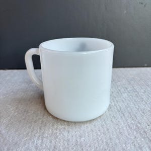 May include: A white ceramic mug with a rounded base and a curved handle. The mug has a smooth, glossy finish and a simple, classic design. The mug is empty and appears clean.