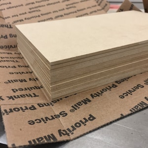 May include: A stack of beige wood panels, likely for crafting or woodworking projects. The panels are neatly stacked and appear to be of uniform size and thickness.
