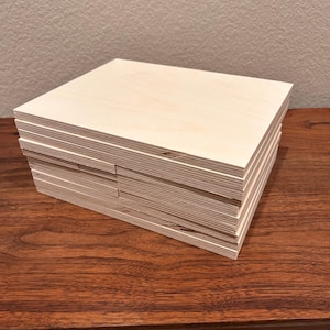 May include: A stack of six unfinished, light brown plywood boards. The boards are stacked neatly on a dark brown wooden surface.