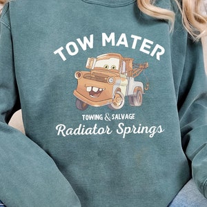 May include: A teal blue sweatshirt with a cartoon image of Tow Mater from the movie Cars. The text "Tow Mater" is printed in white above the image. Below the image, the text "Towing & Salvage Radiator Springs" is printed in white.