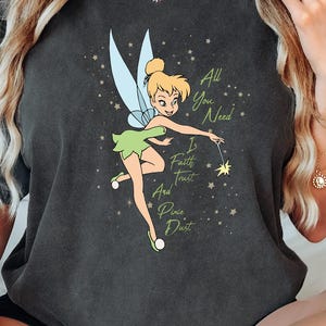 May include: Dark gray t-shirt featuring a cartoon image of Tinkerbell with text that reads "All You Need Is Faith, Trust, And Pixie Dust." The fairy is depicted with blonde hair, a green dress, and blue wings. The shirt has a vintage, washed look.