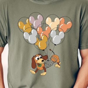 Może przedstawiać: Oliwkowo-zielona koszulka z kresk&oacute;wkową ilustracją Slinky Dog z Toy Story, uniesionego przez grupę balon&oacute;w w kształcie Myszki Miki w r&oacute;żnych kolorach. Projekt jest wyśrodkowany na koszulce.
