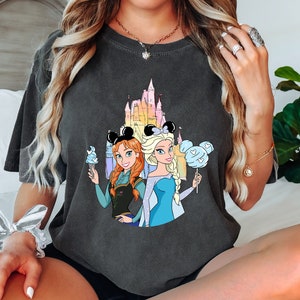 Puede incluir: Una camiseta gris oscuro con un estampado gr&aacute;fico de las princesas Disney Elsa y Anna de Frozen. El gr&aacute;fico muestra a las princesas de pie frente al castillo de Cenicienta con un esquema de color azul y rosa. Elsa sostiene un algod&oacute;n de az&uacute;car azul y Anna sostiene un cono de helado azul.