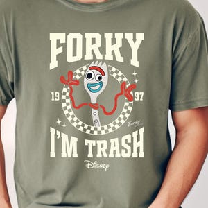 May include: A green t-shirt with a white graphic of the character Forky from the Toy Story movie. The graphic includes the text "Forky I'm Trash" and the Disney logo.