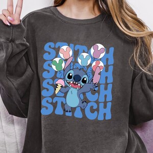 May include: Dark gray sweatshirt featuring a cartoon Stitch character holding ice cream cones. The word "Stitch" is printed in blue, with colorful ice cream cone graphics. The sweatshirt is worn with a pink beanie.