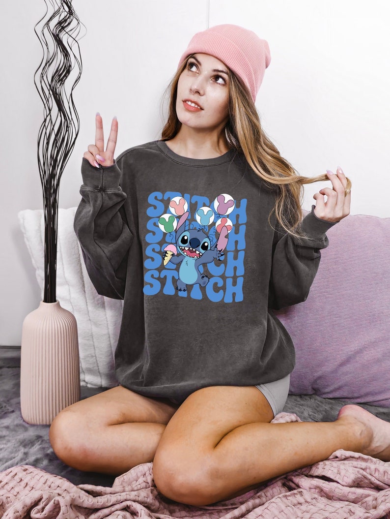 May include: Dark gray sweatshirt featuring a cartoon Stitch character holding ice cream cones. The word "Stitch" is printed in blue, with colorful ice cream cone graphics. The sweatshirt is worn with a pink beanie.