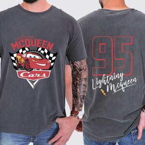 May include: A dark grey t-shirt featuring a red, white and black graphic of Lightning McQueen from the Cars movie. The back of the shirt has the number 95 in red with the text "Lightning McQueen" in white.