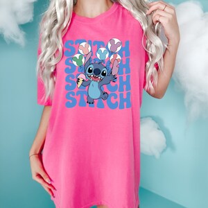 May include: A bright pink t-shirt featuring a cartoon character holding ice cream and balloons. The word "Stitch" is printed in blue, stacked vertically. The character is blue with large ears and a happy expression.