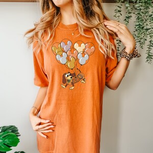 Może przedstawiać: Pomarańczowa koszulka z kresk&oacute;wkową ilustracją Slinky Dog z Toy Story, zawieszonego na grupie balon&oacute;w w kształcie Myszki Miki w r&oacute;żnych kolorach. Koszulka jest w jednolitym pomarańczowym kolorze.