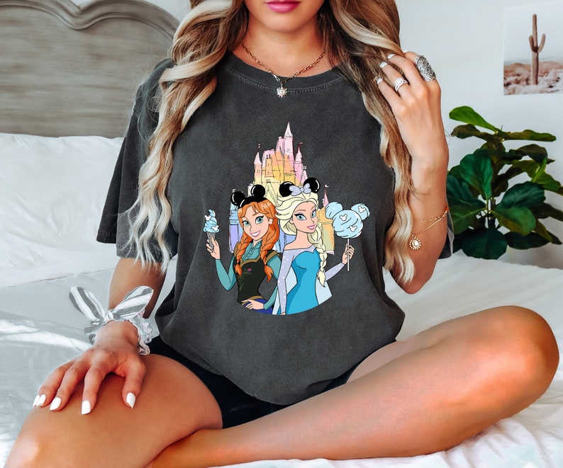Puede incluir: Una camiseta gris oscuro con un estampado gr&aacute;fico de las princesas Disney Elsa y Anna de Frozen. El gr&aacute;fico muestra a las princesas de pie frente al castillo de Cenicienta con un esquema de color azul y rosa. Elsa sostiene un algod&oacute;n de az&uacute;car azul y Anna sostiene un cono de helado azul.