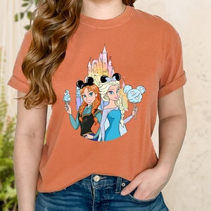 Puede incluir: Una camiseta naranja quemada con un gr&aacute;fico del castillo de Disney, Anna y Elsa de Frozen, con orejas de Mickey Mouse y sosteniendo bocadillos.