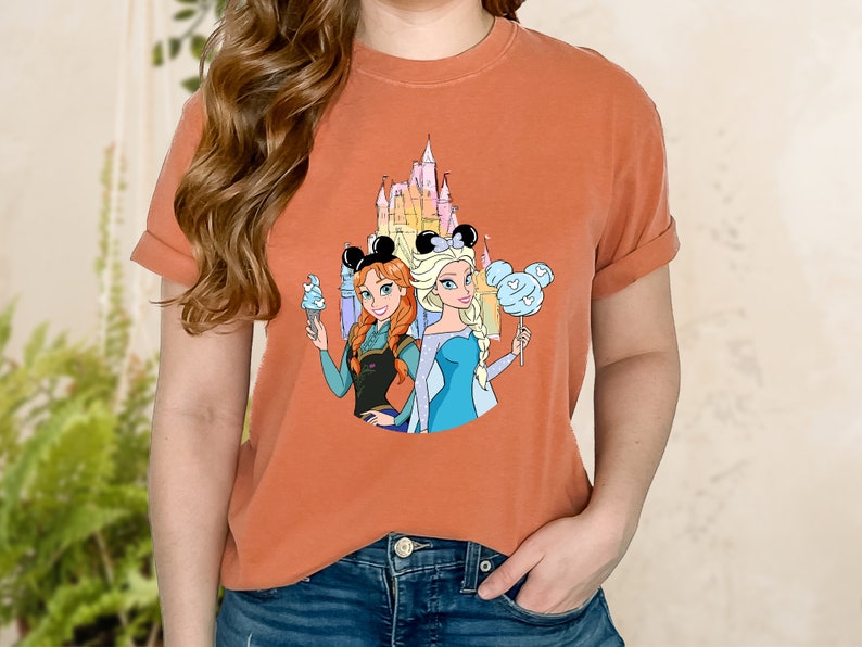 Puede incluir: Una camiseta naranja quemada con un gr&aacute;fico del castillo de Disney, Anna y Elsa de Frozen, con orejas de Mickey Mouse y sosteniendo bocadillos.