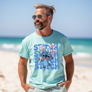 May include: A light blue t-shirt featuring the character Stitch from Lilo & Stitch. Stitch is holding ice cream and balloons. The word "STITCH" is repeated in a blue font. The shirt is short-sleeved.