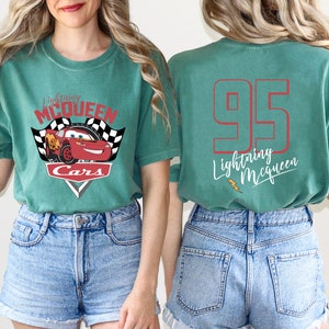 May include: A teal green t-shirt featuring a red and white graphic of Lightning McQueen from the Cars film. The back of the shirt has the number 95 and the text "Lightning McQueen" in white.