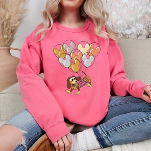Może przedstawiać: R&oacute;żowa bluza z kresk&oacute;wkową ilustracją Slinky Dog z Toy Story, niesionego przez balony w kształcie Myszki Miki w r&oacute;żnych kolorach. Bluza jest w jednolitym r&oacute;żowym kolorze.