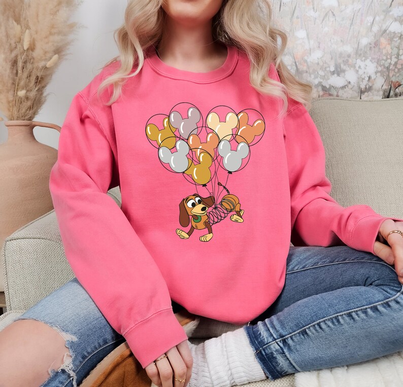 Może przedstawiać: R&oacute;żowa bluza z kresk&oacute;wkową ilustracją Slinky Dog z Toy Story, niesionego przez balony w kształcie Myszki Miki w r&oacute;żnych kolorach. Bluza jest w jednolitym r&oacute;żowym kolorze.