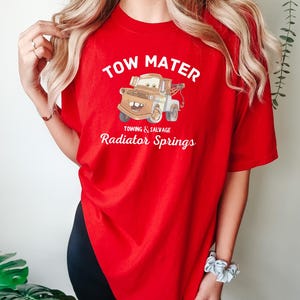 May include: Red t-shirt with a cartoon image of Tow Mater from the Cars movie. The text "Tow Mater" is printed in white above the image. Below the image, the text "Towing & Salvage Radiator Springs" is printed in white.