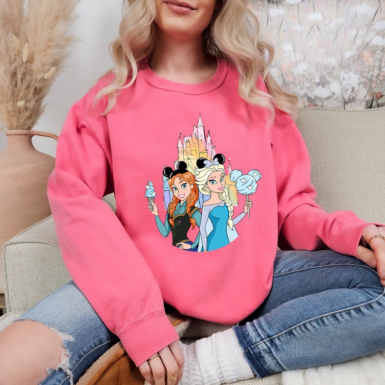 Puede incluir: Sudadera rosa con un estampado gr&aacute;fico del castillo de Disney, Elsa y Anna de Frozen con orejas de rat&oacute;n y sosteniendo helado y algod&oacute;n de az&uacute;car.