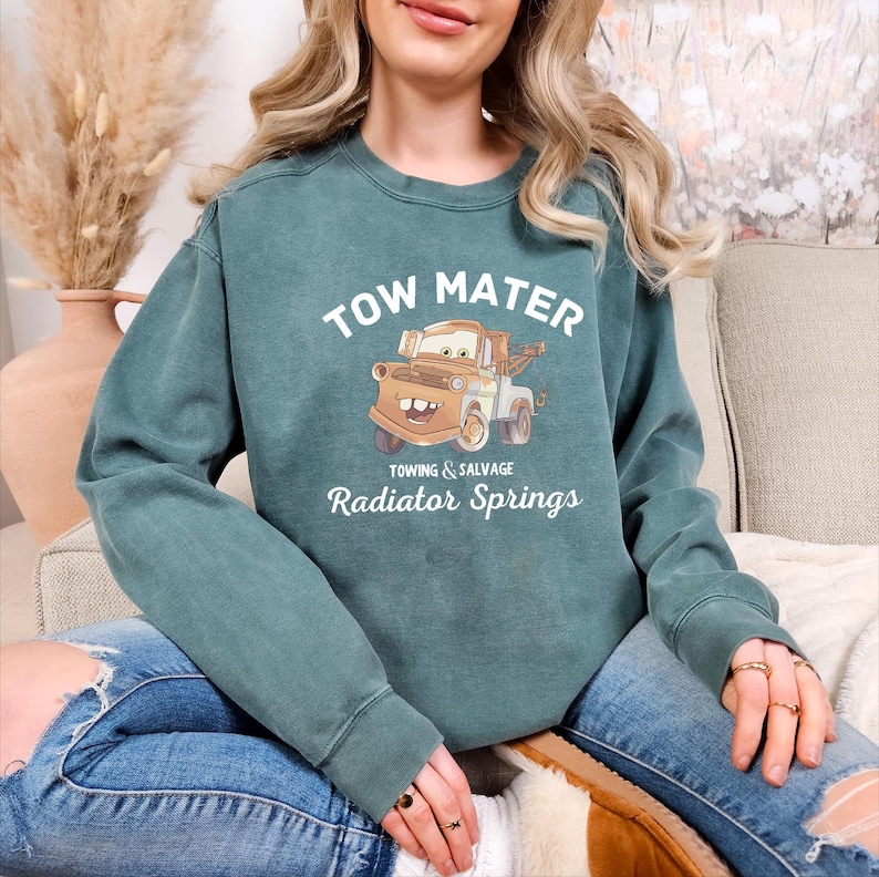 May include: A teal blue sweatshirt with a cartoon image of Tow Mater from the movie Cars. The text "Tow Mater" is printed in white above the image. Below the image, the text "Towing & Salvage Radiator Springs" is printed in white.