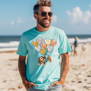 Może przedstawiać: Jasnoniebieska koszulka z wizerunkiem kresk&oacute;wkowego psa trzymającego balony w kształcie uszu Myszki Miki. Balony są w r&oacute;żnych kolorach, w tym ż&oacute;łtym, pomarańczowym i szarym. Koszulka wykonana jest z miękkiego materiału.