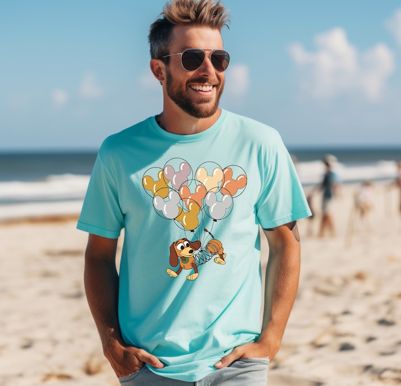 Może przedstawiać: Jasnoniebieska koszulka z wizerunkiem kresk&oacute;wkowego psa trzymającego balony w kształcie uszu Myszki Miki. Balony są w r&oacute;żnych kolorach, w tym ż&oacute;łtym, pomarańczowym i szarym. Koszulka wykonana jest z miękkiego materiału.