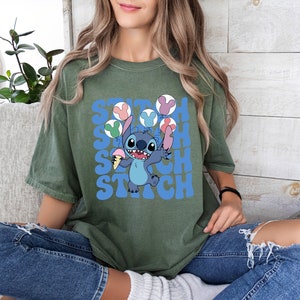 May include: Olive green t-shirt featuring a cartoon Stitch character holding ice cream and balloons. The word "Stitch" is printed in blue, stacked three times. The shirt is a relaxed fit.