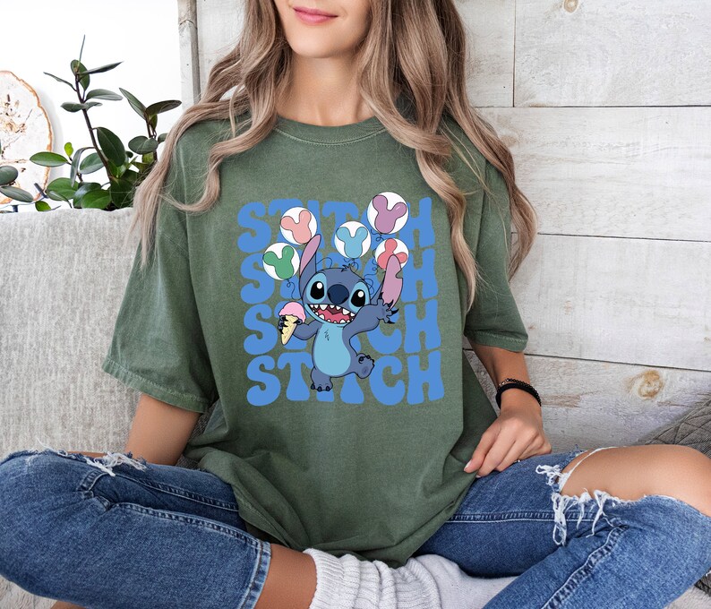 May include: Olive green t-shirt featuring a cartoon Stitch character holding ice cream and balloons. The word "Stitch" is printed in blue, stacked three times. The shirt is a relaxed fit.