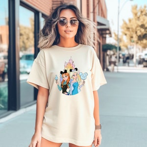 Puede incluir: Una camiseta de color beige claro con un estampado gr&aacute;fico de dos princesas de dibujos animados, Anna y Elsa, que llevan orejas de Mickey Mouse y sostienen conos de helado y algod&oacute;n de az&uacute;car. El gr&aacute;fico tambi&eacute;n incluye una imagen de dibujos animados del castillo de Cenicienta.