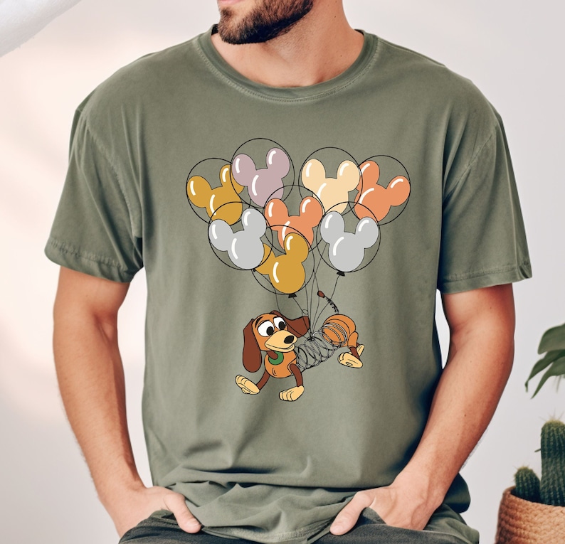 Może przedstawiać: Oliwkowo-zielona koszulka z kresk&oacute;wkową ilustracją Slinky Dog z Toy Story, uniesionego przez grupę balon&oacute;w w kształcie Myszki Miki w r&oacute;żnych kolorach. Projekt jest wyśrodkowany na koszulce.