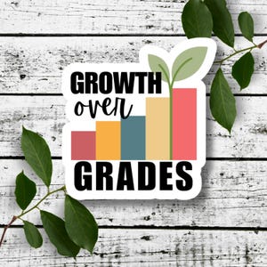 May include: A sticker with the words "GROWTH over GRADES" in black, above a colorful bar graph design. The graph has bars in red, orange, blue, and yellow, with a green sprout at the top. The sticker is on a white wooden background.