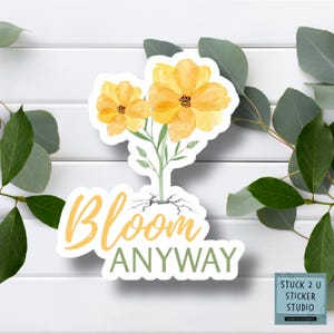 May include: A sticker featuring two yellow watercolor flowers with green stems and the words "Bloom Anyway" in yellow and green. The sticker has a white border and is surrounded by green leaves.