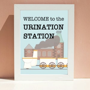 Puede incluir: Una impresión enmarcada en blanco con el texto "WELCOME to the URINATION STATION" y una ilustración de dibujos animados de un tren de inodoros. El tren es azul, blanco y marrón con humo negro saliendo de la chimenea.
