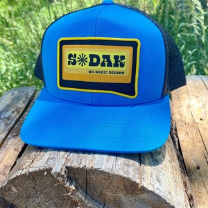 May include: A blue and black trucker hat with a yellow and black patch that says "S*DAK OH GEEZ! DESIGN".