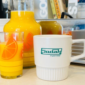 May include: A white ceramic mug with a green logo that says "Sodak! oh geez! design". The mug is sitting on a table next to a glass jug and glass of orange juice.