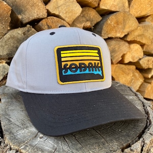 May include: A gray and black baseball cap with a yellow, orange, blue, and black patch that says "SODAK" on it. The patch has a sunset design with three horizontal stripes.