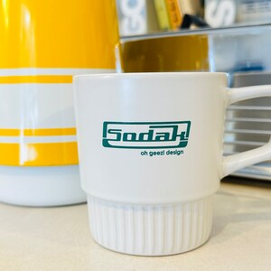 May include: White ceramic mug with a green logo that says "Sodak! oh geezl design".
