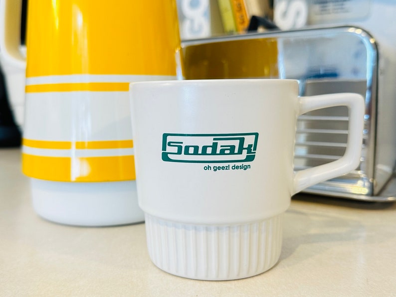 May include: White ceramic mug with a green logo that says "Sodak! oh geezl design".