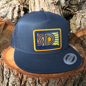 May include: Navy blue trucker hat with a mesh back and a flat brim. The front features a rectangular patch with the letters "SD" in orange and yellow, with a sunburst design. The hat is sitting on a wooden surface.