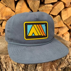 May include: A dark blue trucker hat with a yellow, orange, and blue patch featuring a stylized tent design. The patch has a black outline and a yellow border.